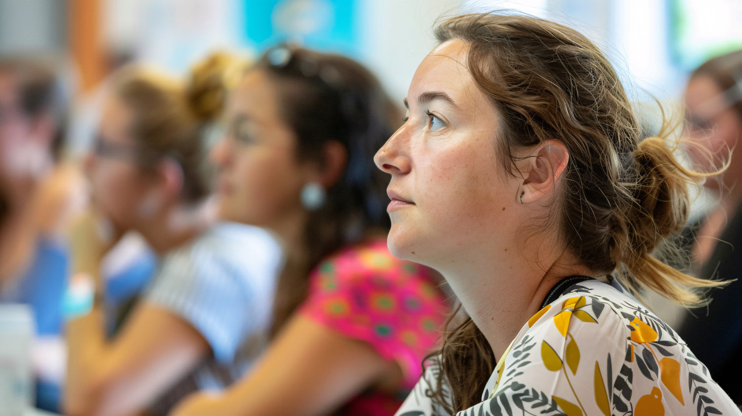 A white teacher with long brown hair sits in a workshop and is paying attention looking towards the speaker.